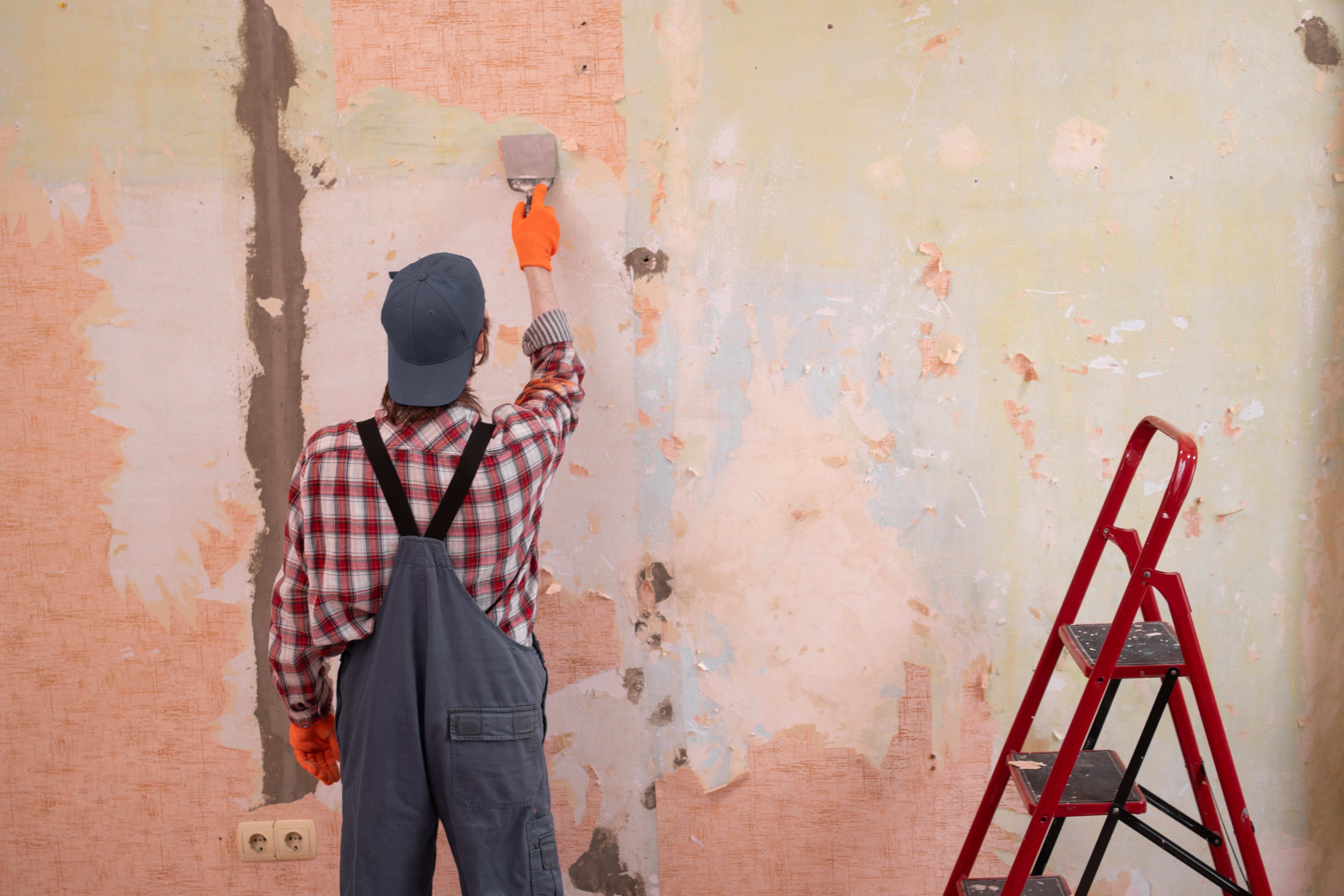 Professional repairman stripping old wallpaper from wall with metal spatula for apartment renovation. Home renovation and house renewal concept.